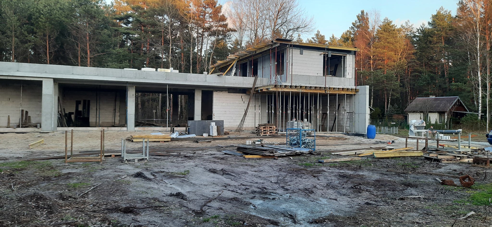 Budowa domu jednorodzinnego w stanie surowym otwartym, widoczne ściany z bloczków, szalunki stropowe wsparte na stemplach, teren budowy z materiałami i narzędziami, w tle las iglasty i mały budynek...
