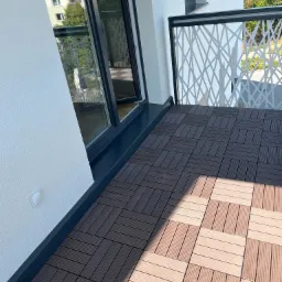 Nowoczesny balkon z brązową podłogą z paneli kompozytowych ułożonych w kwadraty, biała balustrada z ażurowym wzorem geometrycznym i widok na budynek mieszkalny w tle.