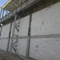 Wznoszenie ściany z białych bloczków betonowych, widoczna stalowa konstrukcja szkieletowa budynku i zbrojenie pionowe w betonie.