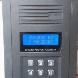 Szary panel domofonu marki Elfon z wyświetlaczem LCD, klawiaturą numeryczną i czytnikiem kart zbliżeniowych, zamontowany na ścianie.