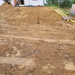 Przygotowany teren pod budowę domu jednorodzinnego, świeżo wyrównana ziemia z widocznymi śladami opon, częściowo ułożone żółte rury, w tle fragment budynku z otwartymi drzwiami i pracownikiem...