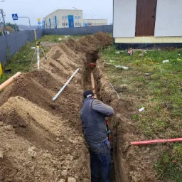 Pracownik w wykopie, montuje rury kanalizacyjne w pobliżu budynku. Widoczne narzędzia pomiarowe, poziomica i sterta ziemi.