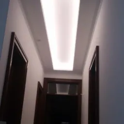 Korytarz z oświetleniem sufitowym w postaci podłużnego panelu LED, widoczne dwie pary drzwi z ciemnego drewna i białe ściany z dekoracyjną listwą przysufitową.