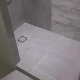 Wykończona kabina prysznicowa z szarymi, kwadratowymi płytkami, czarnym odpływem liniowym i imitacją drewna na jednej ze ścian, widoczna instalacja wodna przed zabudową.