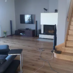 Jasne, drewniane panele podłogowe w salonie z kominkiem i schodami, widoczne gniazdka elektryczne i kable na podłodze.