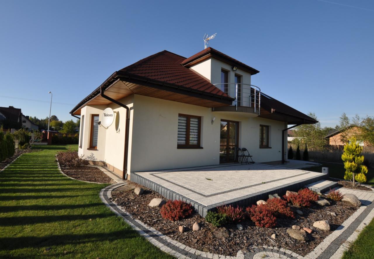 Widok z zewnątrz na parterowy dom z poddaszem, z brązowym dachem, kremową elewacją, balkonem i tarasem wyłożonym jasną kostką brukową, otoczony zadbanym trawnikiem i rabatami z czerwonymi krzewami.