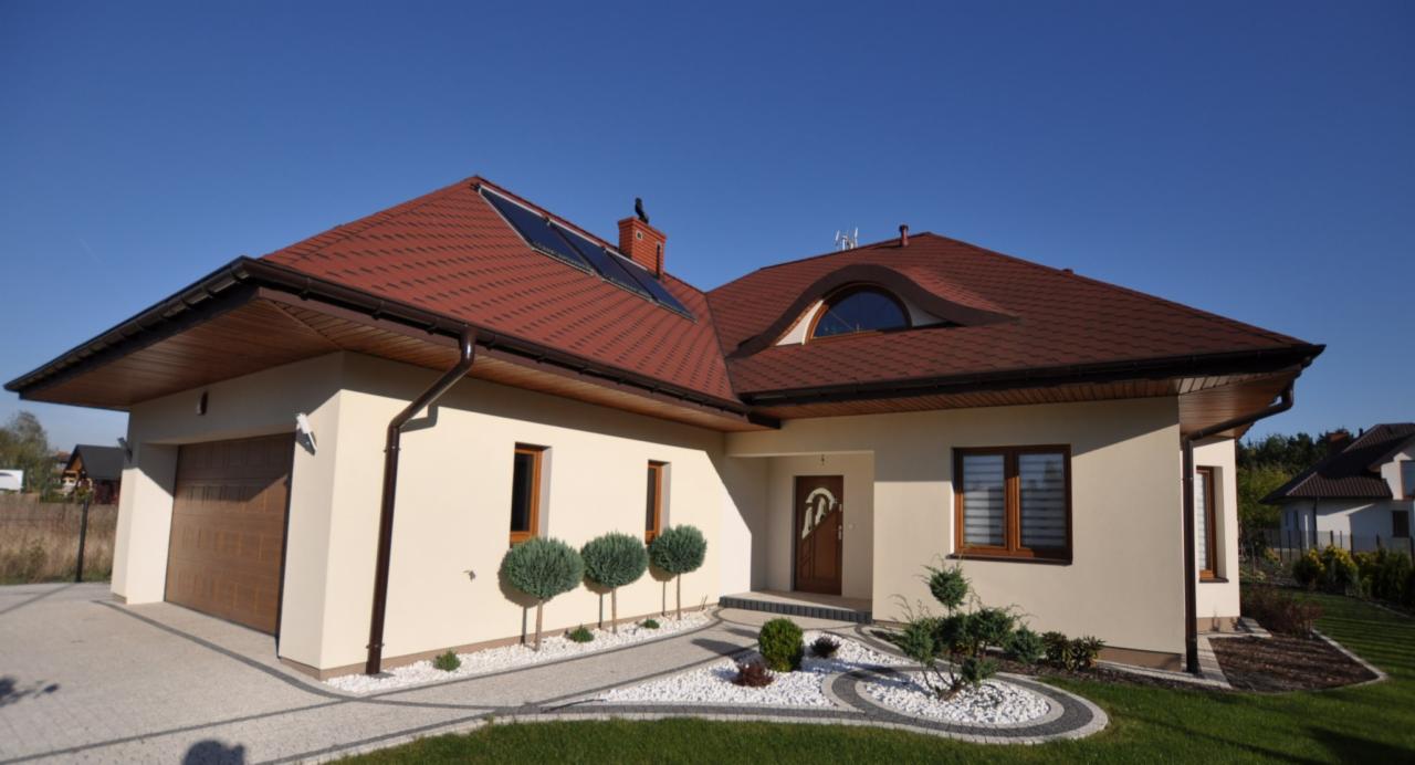 Elegancki dom jednorodzinny z beżową elewacją, czerwoną dachówką, panelami słonecznymi na dachu i zagospodarowanym ogrodem z białymi kamieniami i niskimi drzewkami.
