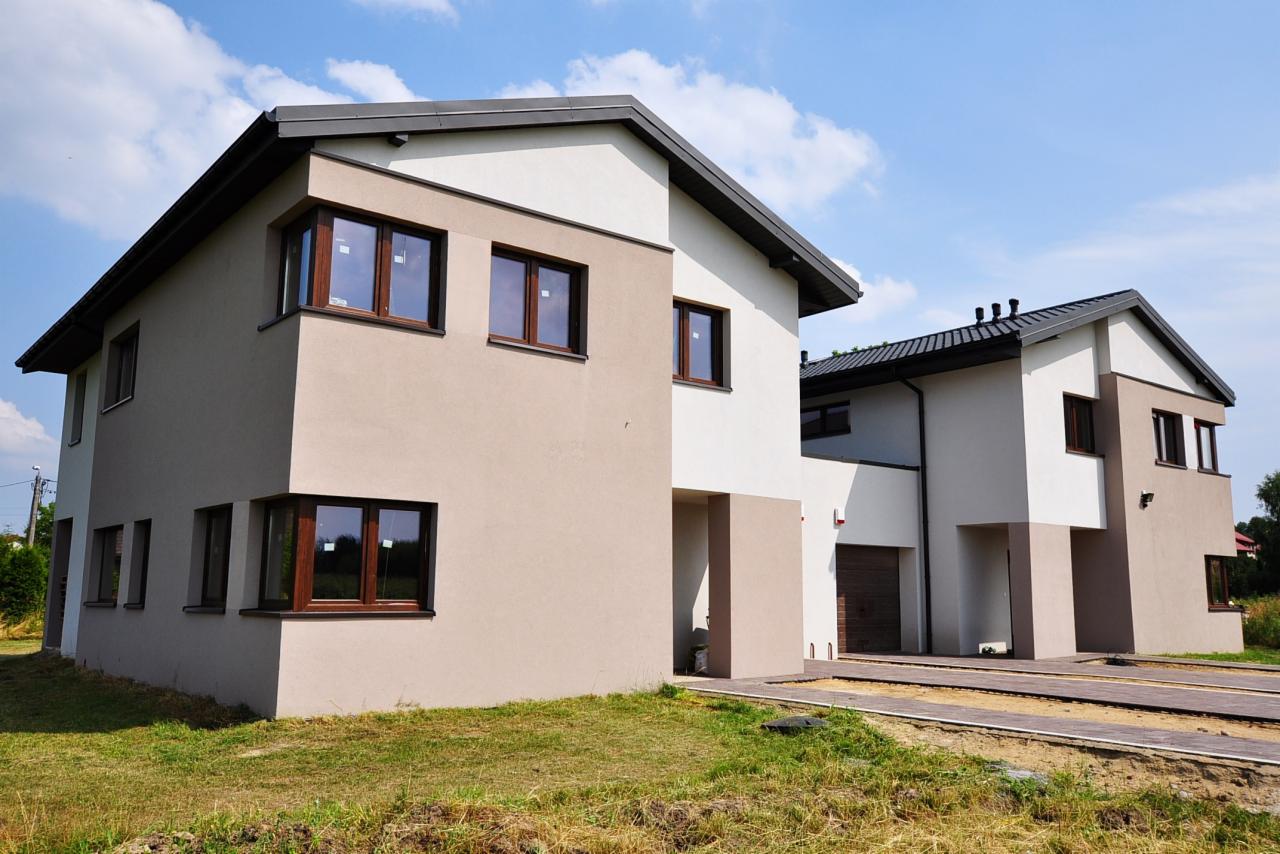 Dwa nowoczesne, dwukondygnacyjne domy w zabudowie bliźniaczej, z jasną elewacją w odcieniach beżu i bieli, brązowe okna, ciemne dachy, trawnik z przodu, błękitne niebo z chmurami w tle.