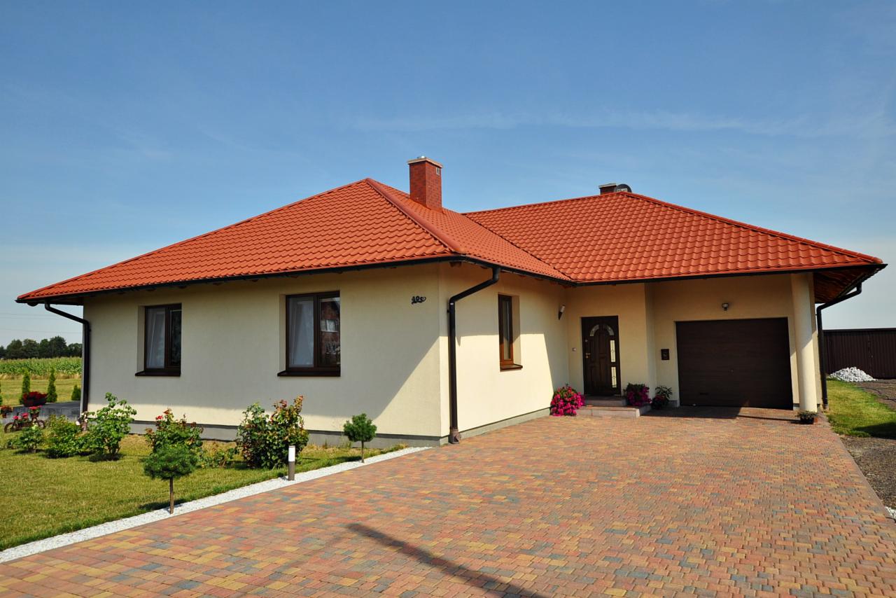 Parterowy dom z czerwoną dachówką, beżową elewacją i garażem w bryle, otoczony niską zielenią i wyłożony kostką brukową w odcieniach czerwieni i brązu.