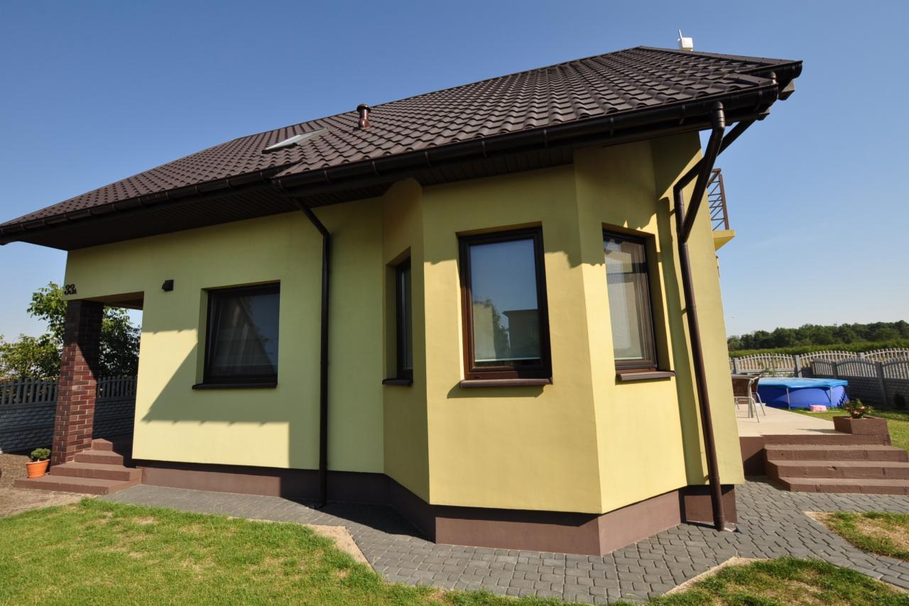 Żółty dom z brązowym dachem i oknami, widoczny fragment ogrodu z basenem w tle.