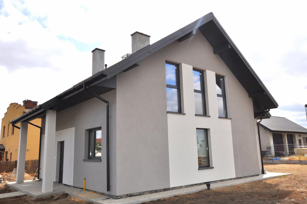 Nowoczesny dom jednorodzinny z szarą elewacją i ciemną stolarką okienną, widok z zewnątrz na fasadę boczną i frontową, z fragmentem niezagospodarowanego terenu.