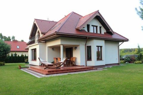 Nowoczesny dom murowany z czerwoną dachówką i tarasem z drewnianymi meblami ogrodowymi, widok z zadbanego trawnika.