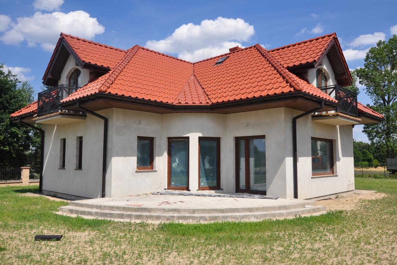 Dwukondygnacyjny dom z czerwonym dachem, balkonami i oknami z brązowymi ramami, w trakcie wykończenia, z widocznym ociepleniem na elewacji i przygotowanym tarasem.
