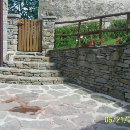 Kamienne schody prowadzące do drewnianej furtki, otoczone kamienną ścianą z drewnianym ogrodzeniem i kwiatami w donicach, na tle kamiennego budynku.