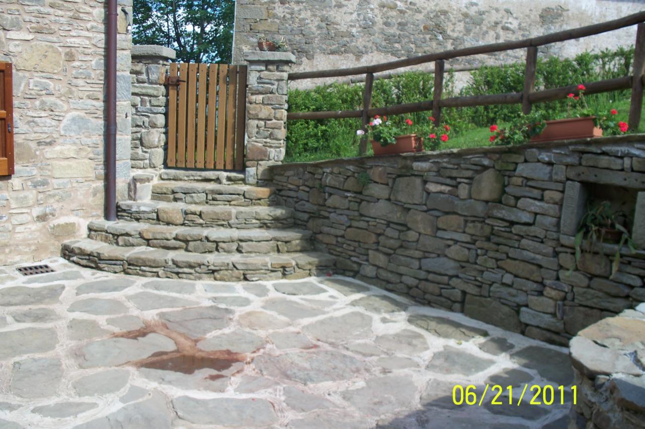 Kamienne schody prowadzące do drewnianej furtki, otoczone kamienną ścianą z drewnianym ogrodzeniem i kwiatami w donicach, na tle kamiennego budynku.