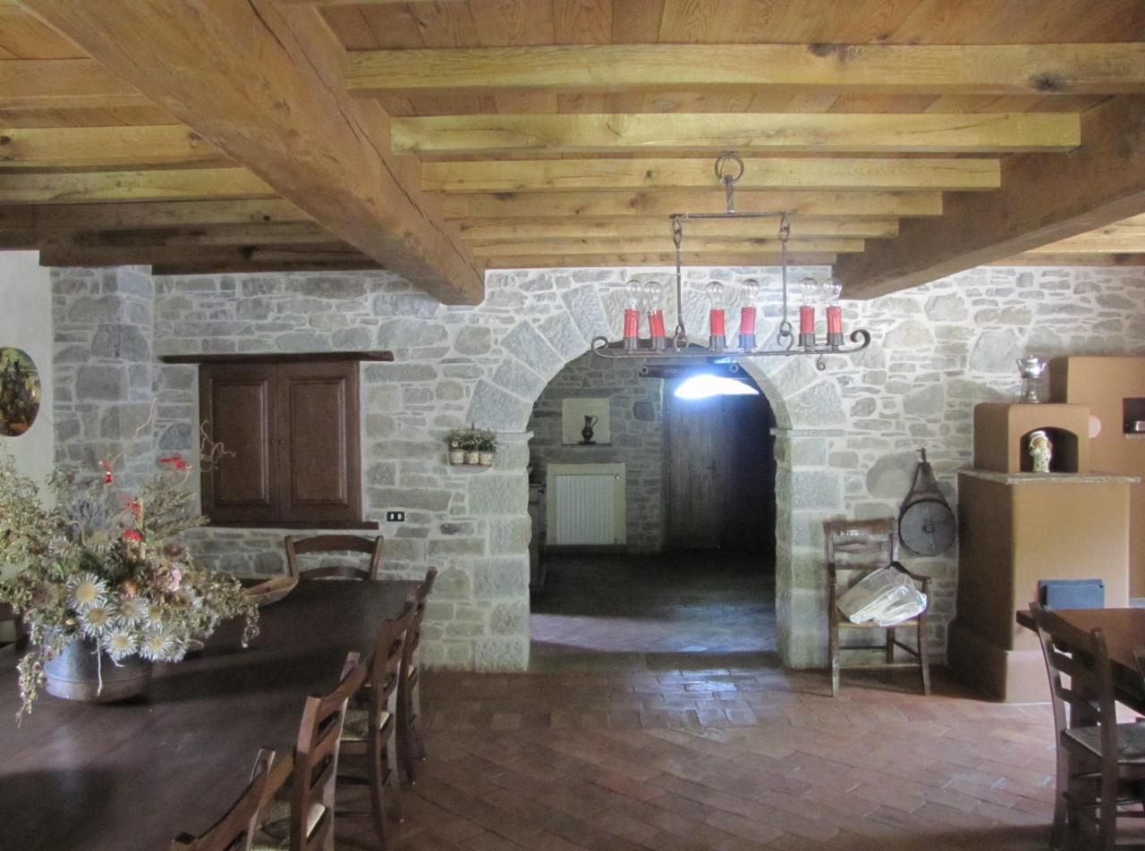 Rustykalne wnętrze z kamiennymi ścianami, drewnianym stropem belkowym i ceglaną podłogą w jodełkę, oświetlone żyrandolem z żarówkami w kształcie świec.