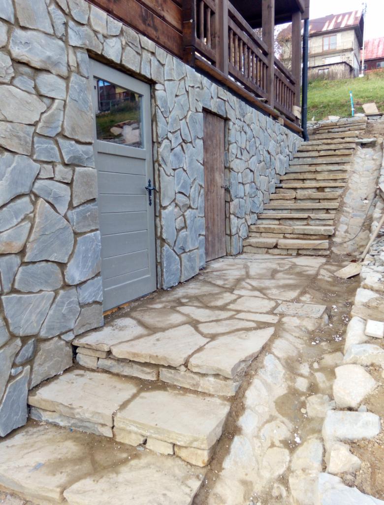 Kamienna elewacja budynku z drewnianym balkonem i kamiennymi schodami w Zamościu, widok na szarą drzwi i drewniane drzwi, teren w trakcie prac wykończeniowych.