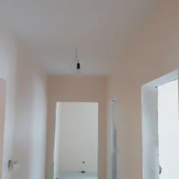 Korytarz po odświeżeniu ścian w odcieniach beżu, widoczne surowe ościeżnice i żarówka zwisająca na kablu z sufitu.