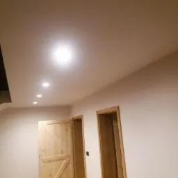 Wnętrze po remoncie: świeżo pomalowane ściany w odcieniach bieli, drewniane drzwi przesuwne w stylu rustykalnym oraz widoczne oświetlenie sufitowe.