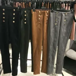 Rząd spodni damskich z ozdobnymi złotymi guzikami w kolorach czarnym, oliwkowym, brązowym i szarym, wiszących na wieszakach w sklepie w Kamienicy Polskiej.