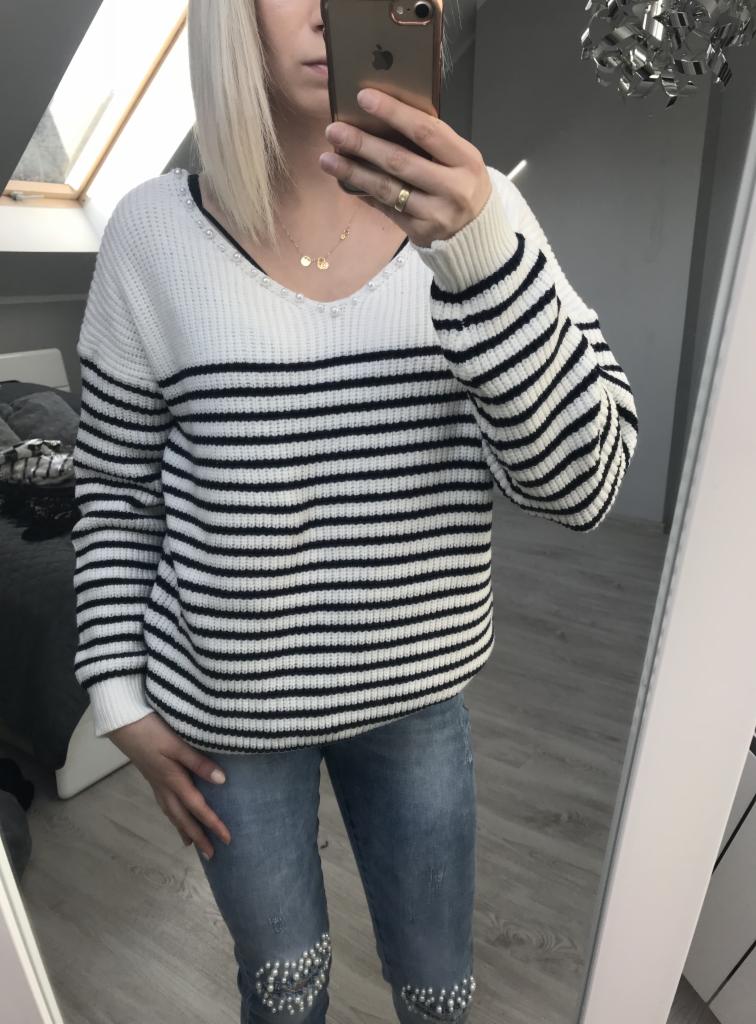 Osoba robi selfie w lustrze, prezentując biało-czarny sweter w paski z perłami przy dekolcie i jeansy z aplikacjami z pereł na kolanach.