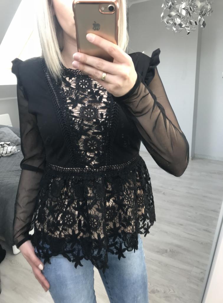 Kobieta robi selfie w lustrze, ubrana w czarną koronkową bluzkę z długimi, prześwitującymi rękawami i dżinsy. Widoczny fragment sypialni z szarym łóżkiem i nowoczesna lampa sufitowa.