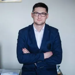 Portret mężczyzny w okularach, ubrany w białą koszulę i granatową marynarkę, pozującego z założonymi rękami na tle jasnej ściany.