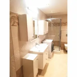 Jasna łazienka z beżowymi płytkami o wzorzystej fakturze, białymi szafkami pod umywalką i wiszącymi, kabiną prysznicową z przezroczystego szkła oraz pralką.