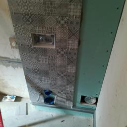 Firma handlowo-usługowa Michał Pytlowany - Montaż płytek z geometrycznym wzorem wokół wnęki na stelaż podtynkowy WC, widoczne niebieskie rury instalacyjne, płyta kartonowo-gipsowa na ścianie obok, czerwona poziomica oparta o ścianę.