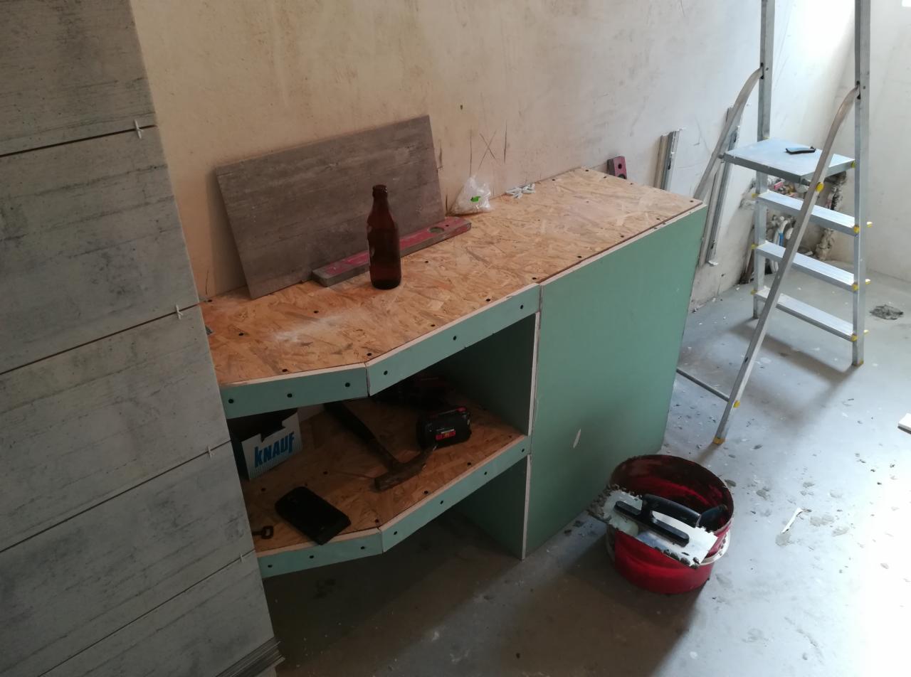 Konstrukcja z płyty karton gips i płyty OSB w trakcie budowy, widoczne narzędzia, poziomica, butelka, wiadro z kielnią zębatą i drabina aluminiowa.