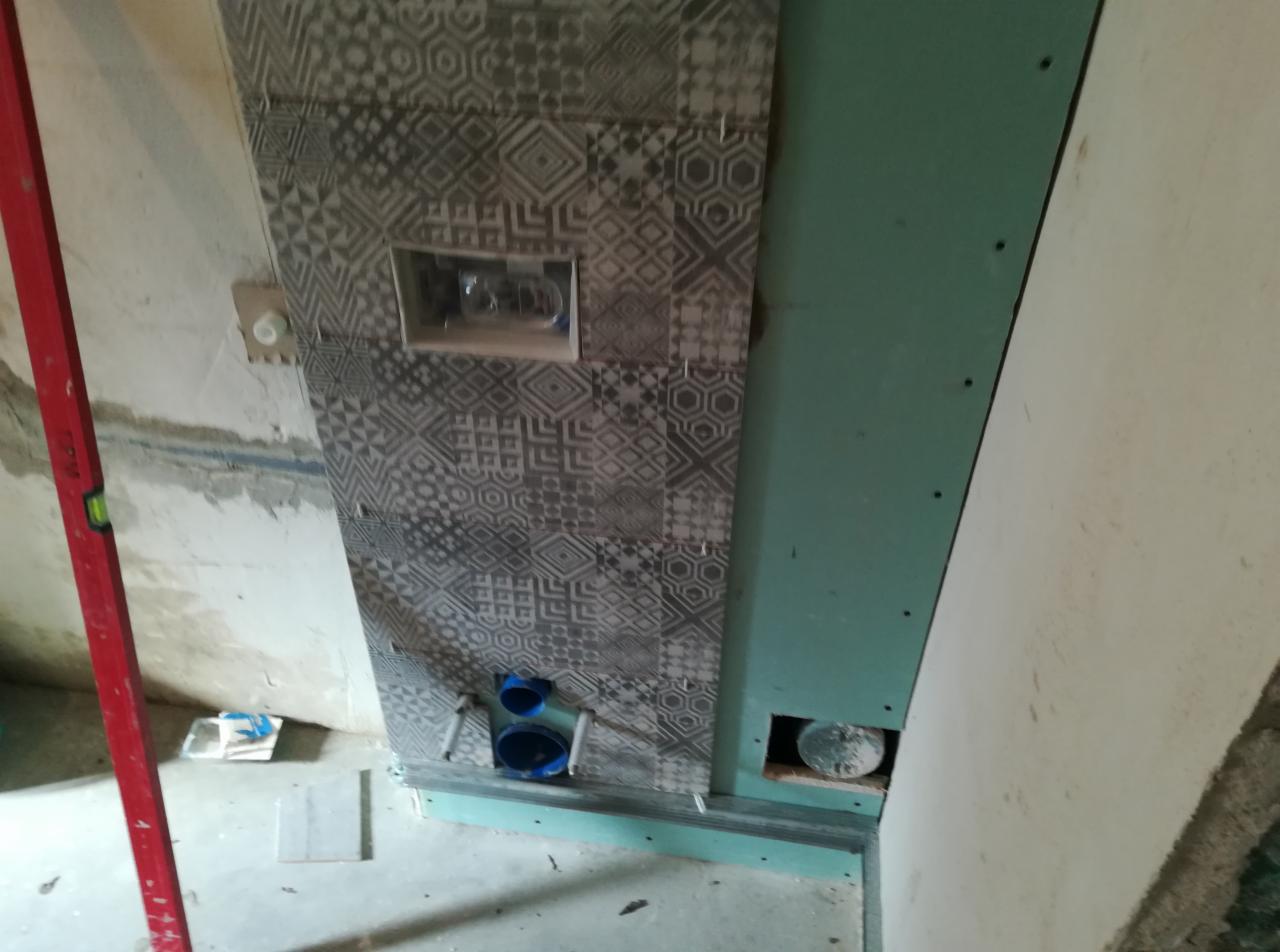 Montaż płytek z geometrycznym wzorem wokół wnęki na stelaż podtynkowy WC, widoczne niebieskie rury instalacyjne, płyta kartonowo-gipsowa na ścianie obok, czerwona poziomica oparta o ścianę.