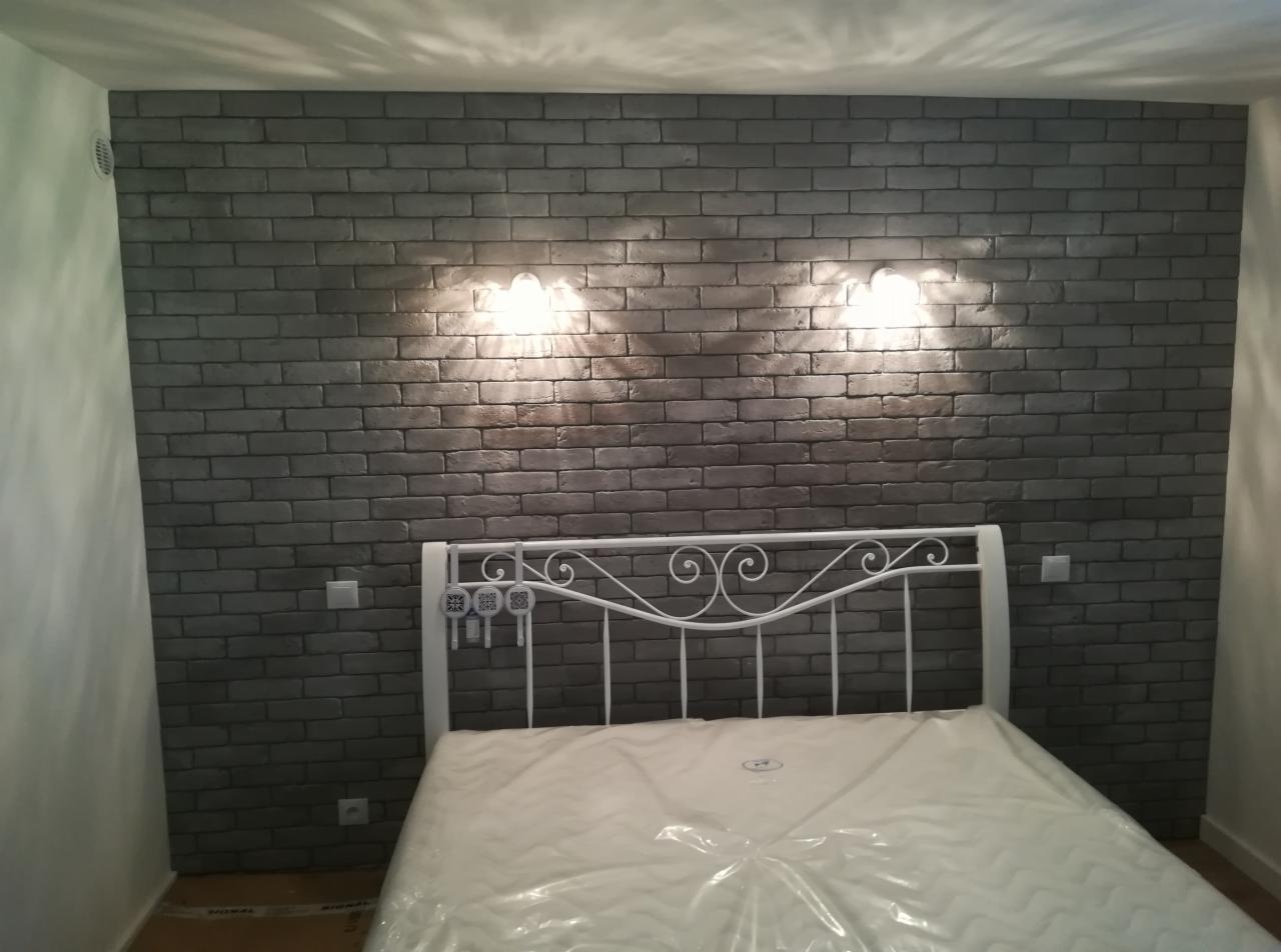 Ściana w sypialni wykończona szarą cegłą dekoracyjną, oświetlona dwoma kinkietami, z widocznym białym łóżkiem z metalowym zagłówkiem i materacem w folii.