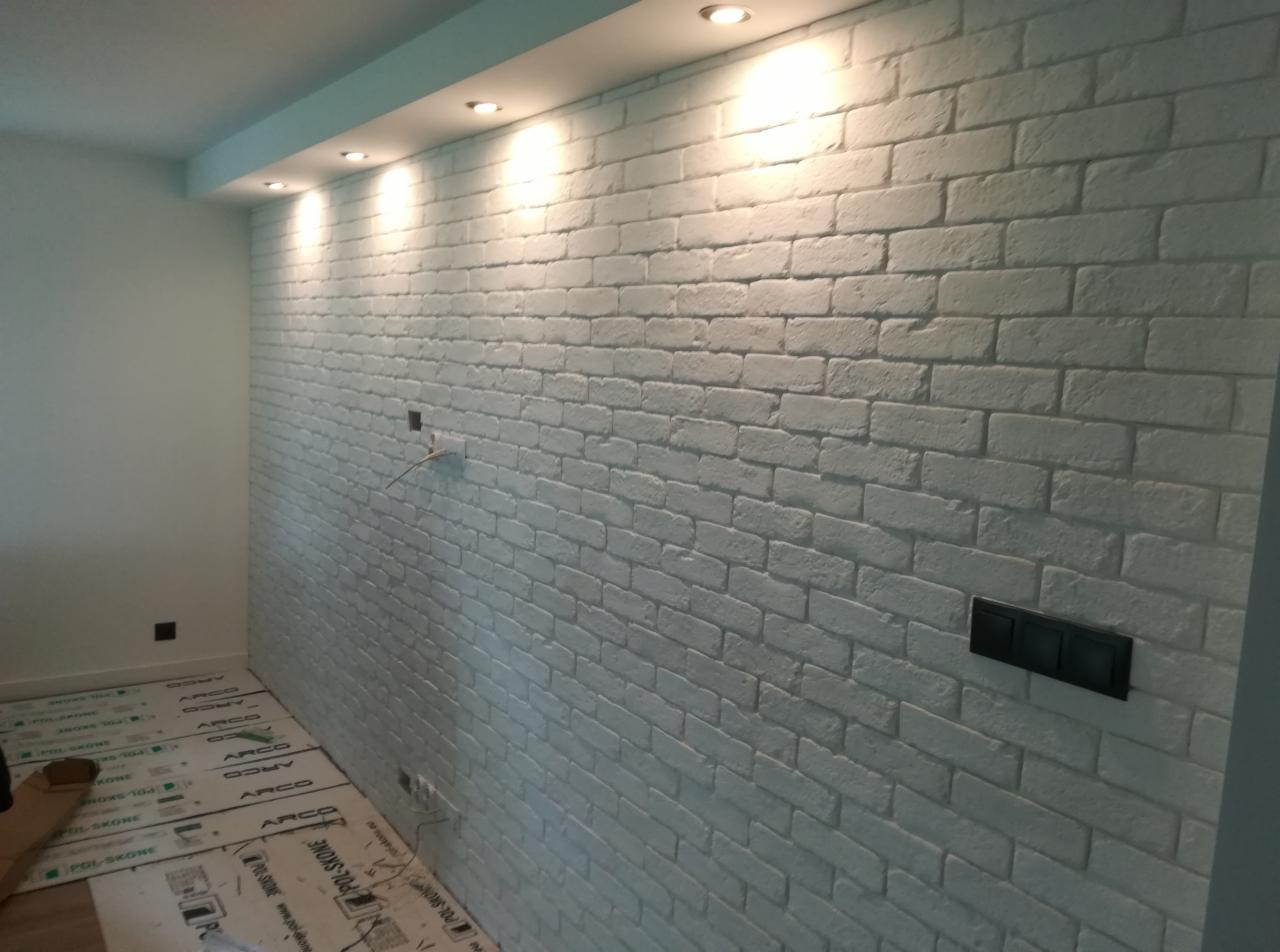 Ściana wewnętrzna wykończona białymi, dekoracyjnymi płytkami imitującymi cegłę, z widocznymi puszkami elektrycznymi i czarnym włącznikiem światła. Na podłodze zabezpieczenie z folii budowlanej...