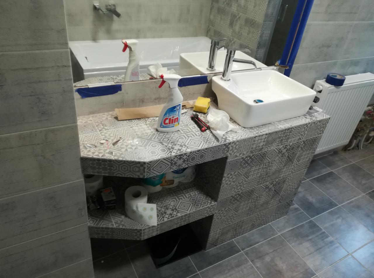 Montaż umywalki nablatowej na szafce z półkami w łazience, widoczne narzędzia i środki czystości, płytki w odcieniach szarości z geometrycznym wzorem.