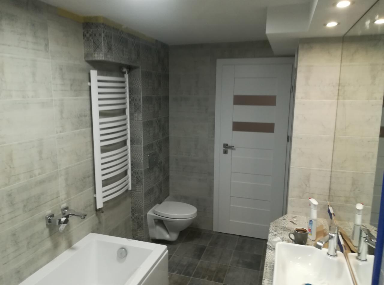 Nowoczesna łazienka w szarościach z grzejnikiem drabinkowym, podwieszaną toaletą i białą armaturą, widok na umywalkę z akcesoriami.