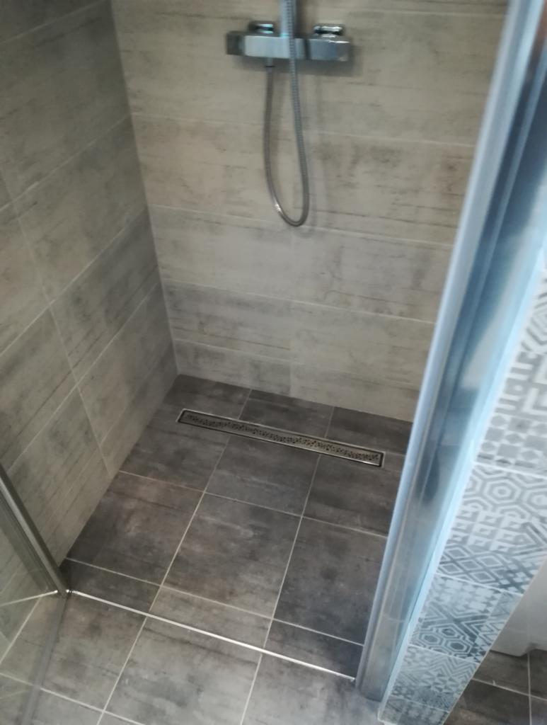 Nowoczesna kabina prysznicowa z odpływem liniowym, wyłożona szarymi płytkami imitującymi kamień, widoczny chromowany zestaw natryskowy.