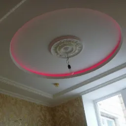 Okrągły, podwieszany sufit z dekoracyjną rozetą i czerwoną obwódką LED, widoczne ozdobne listwy sufitowe i fragment tapety w tle.