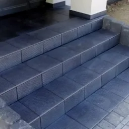 Zewnętrzne schody wejściowe wyłożone ciemnymi, ryflowanymi płytkami, obramowane szarymi betonowymi elementami, z widoczną kostką brukową na podeście.