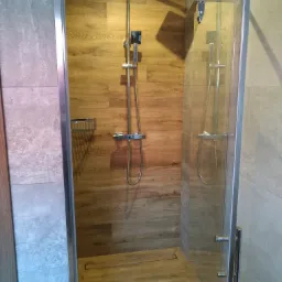 Kabina prysznicowa z deszczownicą i panelem prysznicowym, wykończona płytkami imitującymi drewno i szarym kamieniem, z odpływem liniowym w podłodze.