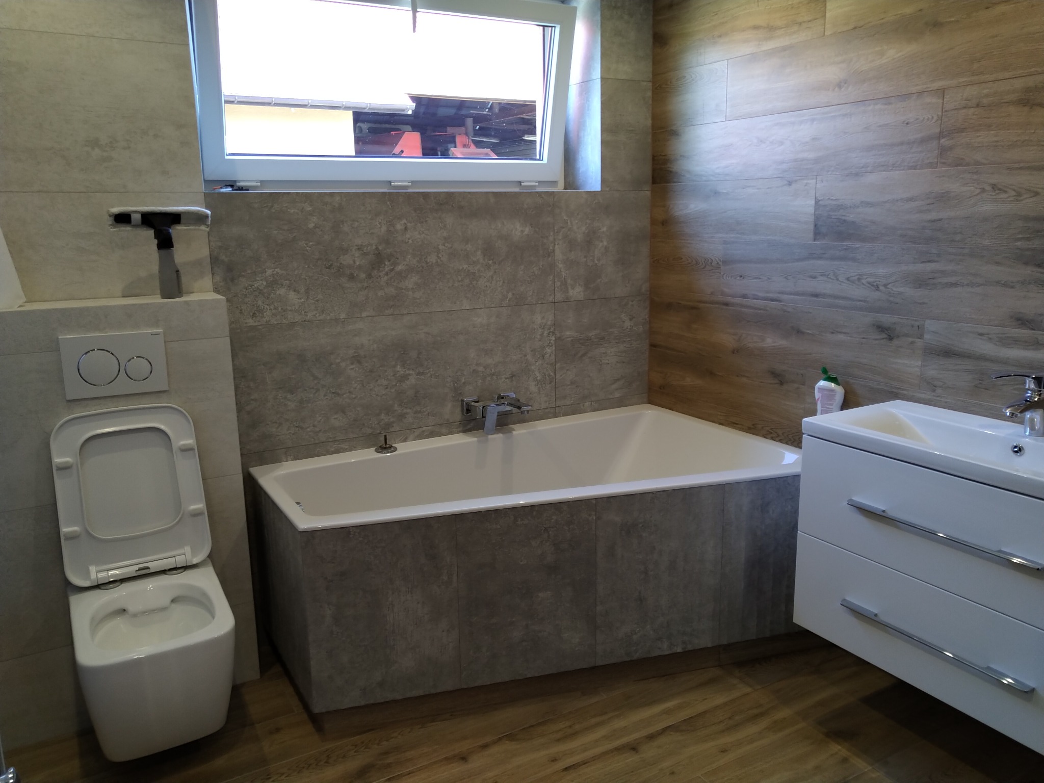 Nowoczesna łazienka z wanną obudowaną szarymi płytkami imitującymi beton, wiszącą toaletą z otwartą klapą, białą szafką z umywalką i drewnopodobnymi płytkami na ścianie po prawej stronie.