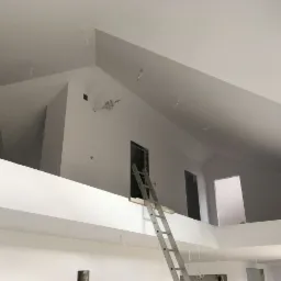 Wnętrze domu po remoncie, świeżo pomalowane na biało, widoczna aluminiowa drabina oparta o ścianę na antresoli, wystające kable oświetleniowe z sufitu, przygotowane do montażu lamp.