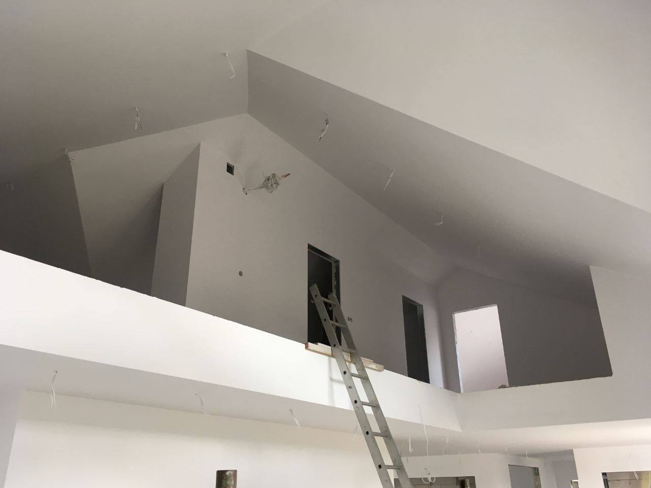 Wnętrze domu po remoncie, świeżo pomalowane na biało, widoczna aluminiowa drabina oparta o ścianę na antresoli, wystające kable oświetleniowe z sufitu, przygotowane do montażu lamp.