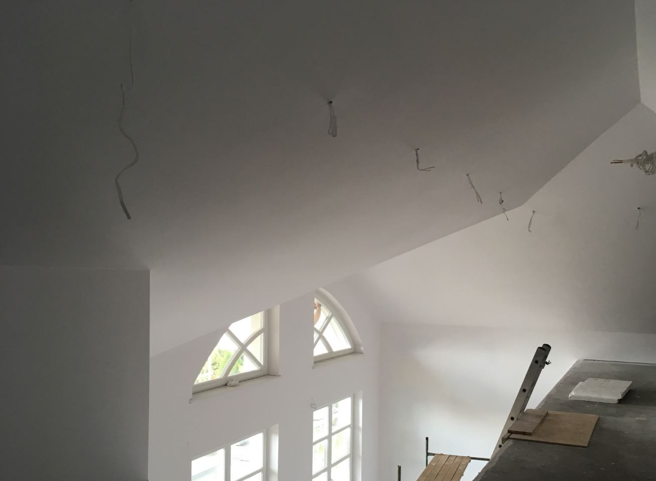 Białe wnętrze w trakcie malowania: widoczne przewody wystające z sufitu, drabina i okna w kształcie łuku.