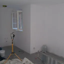 Pokój po malowaniu ścian na biało, widoczna drabina, lampa halogenowa na statywie, wiadro z farbą i otwarte okno z białymi ramami.