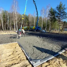 Wylewanie fundamentów pod dom jednorodzinny; widoczna stal zbrojeniowa, szalunki, betonowa pompa z mieszalnikiem w tle oraz dwóch robotników wygładzających powierzchnię betonu.