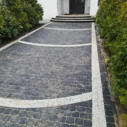 Ułożona kostka brukowa w Zbicznie z białymi, dekoracyjnymi pasami prowadząca do wejścia do budynku, otoczona tujami.