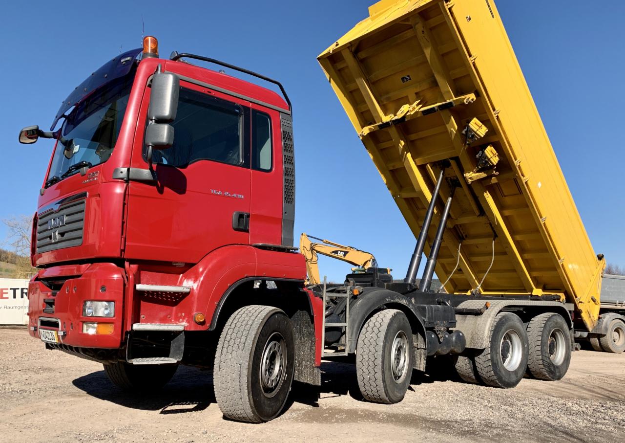 Czerwona ciężarówka MAN z żółtą, podniesioną skrzynią ładunkową, widoczny mały, żółty dźwig na platformie, na tle błękitnego nieba.