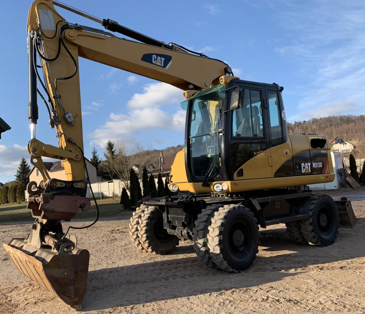 Żółta koparka kołowa CAT M313D stoi na piaszczystym terenie w słoneczny dzień, z widocznym łyżką i ramieniem gotowym do pracy, w tle domy i wzgórze.