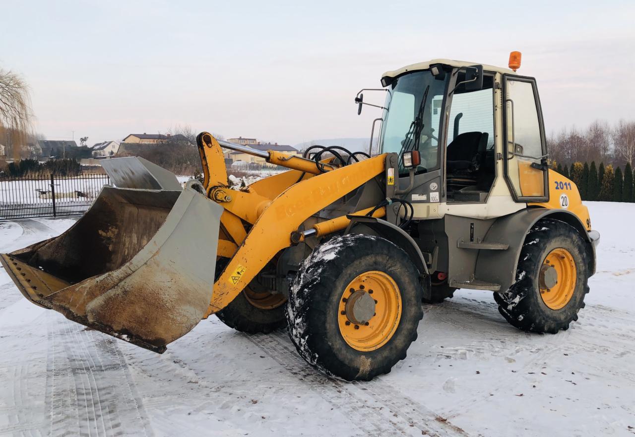 Żółto-szara ładowarka kołowa Liebherr 2011 na śniegu, z widocznymi śladami użytkowania na łyżce, zaparkowana na placu budowy w zimowy dzień.