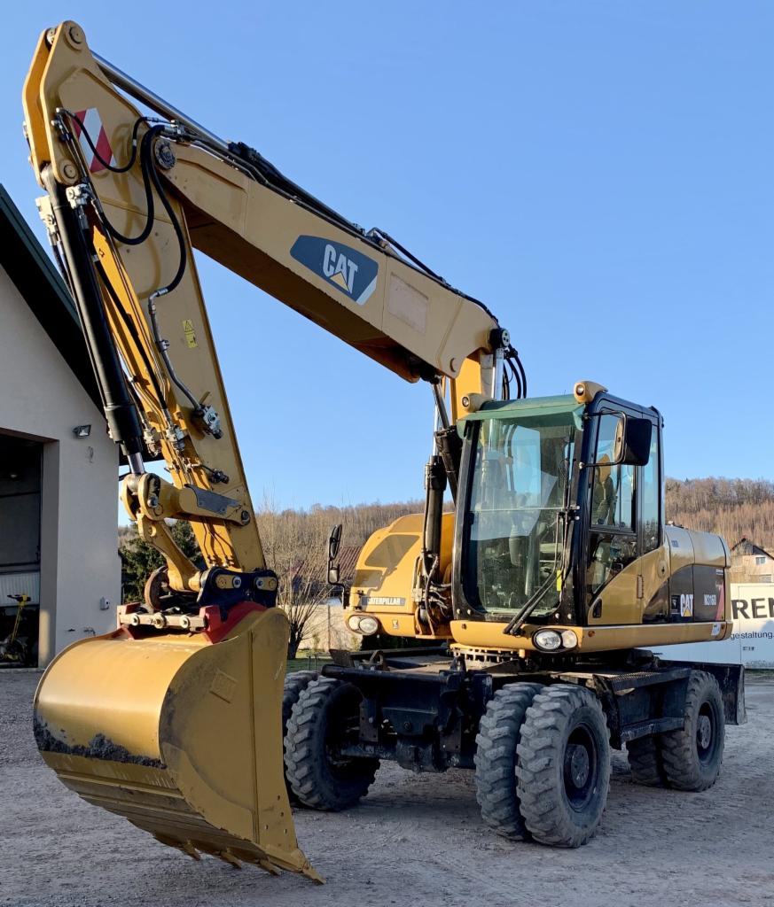 Żółta koparka kołowa CAT M316D w zbliżeniu, widoczna na tle budynku i fragmentu innej maszyny budowlanej, z uniesionym ramieniem i łyżką skierowaną w dół.